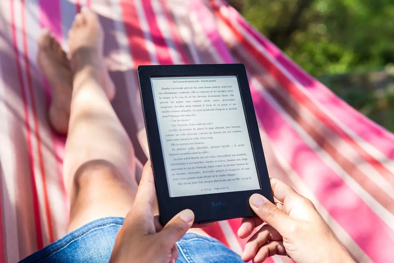 A person reading an ebook on a Kindle while relaxing on a hammock outdoors, with a blurred background of greenery and pink-striped fabric.